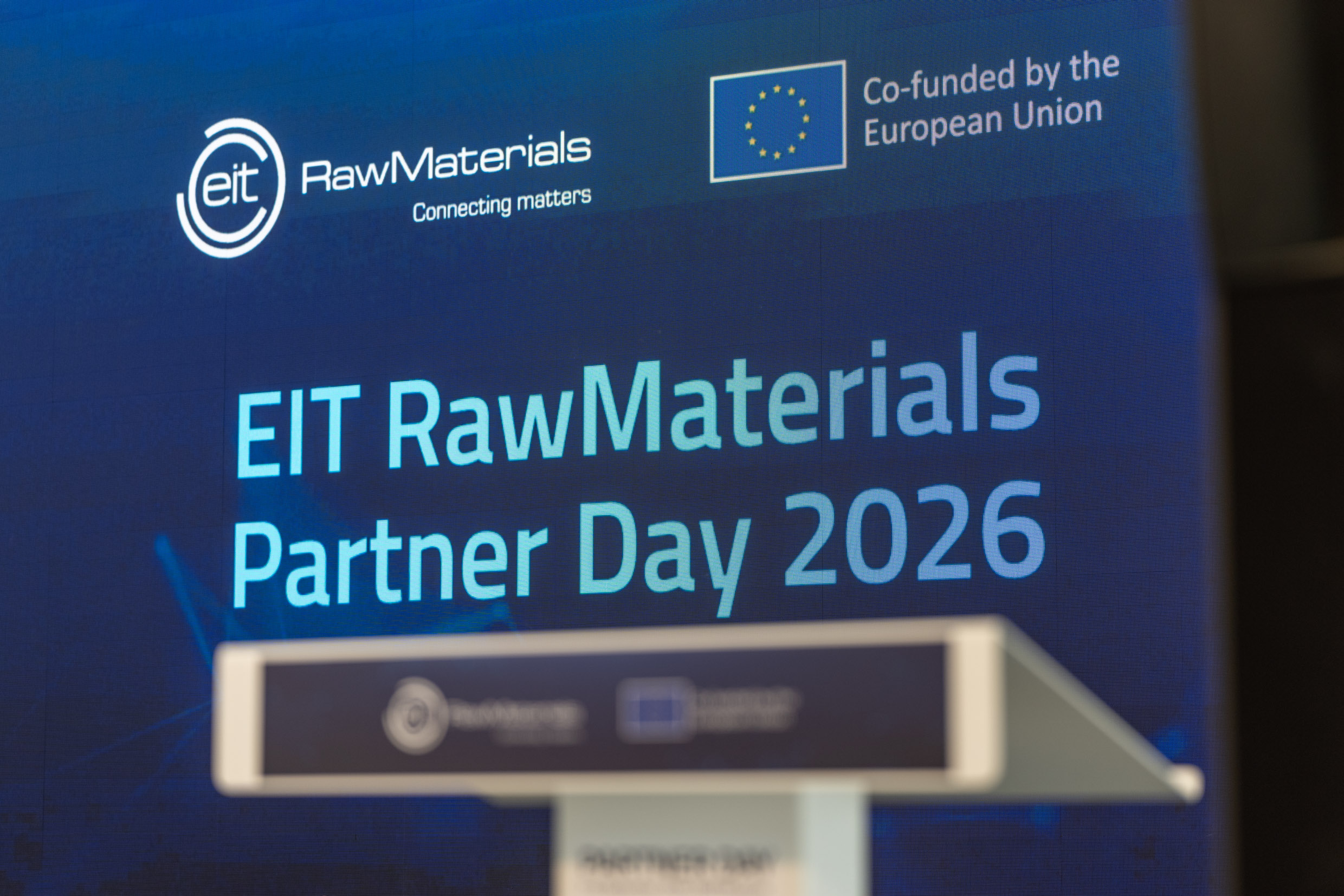White podium in soft focus in front of a large blue digital screen reading "EIT RawMaterials Partner Day 2026" with the EIT RawMaterials and European Union logos above.