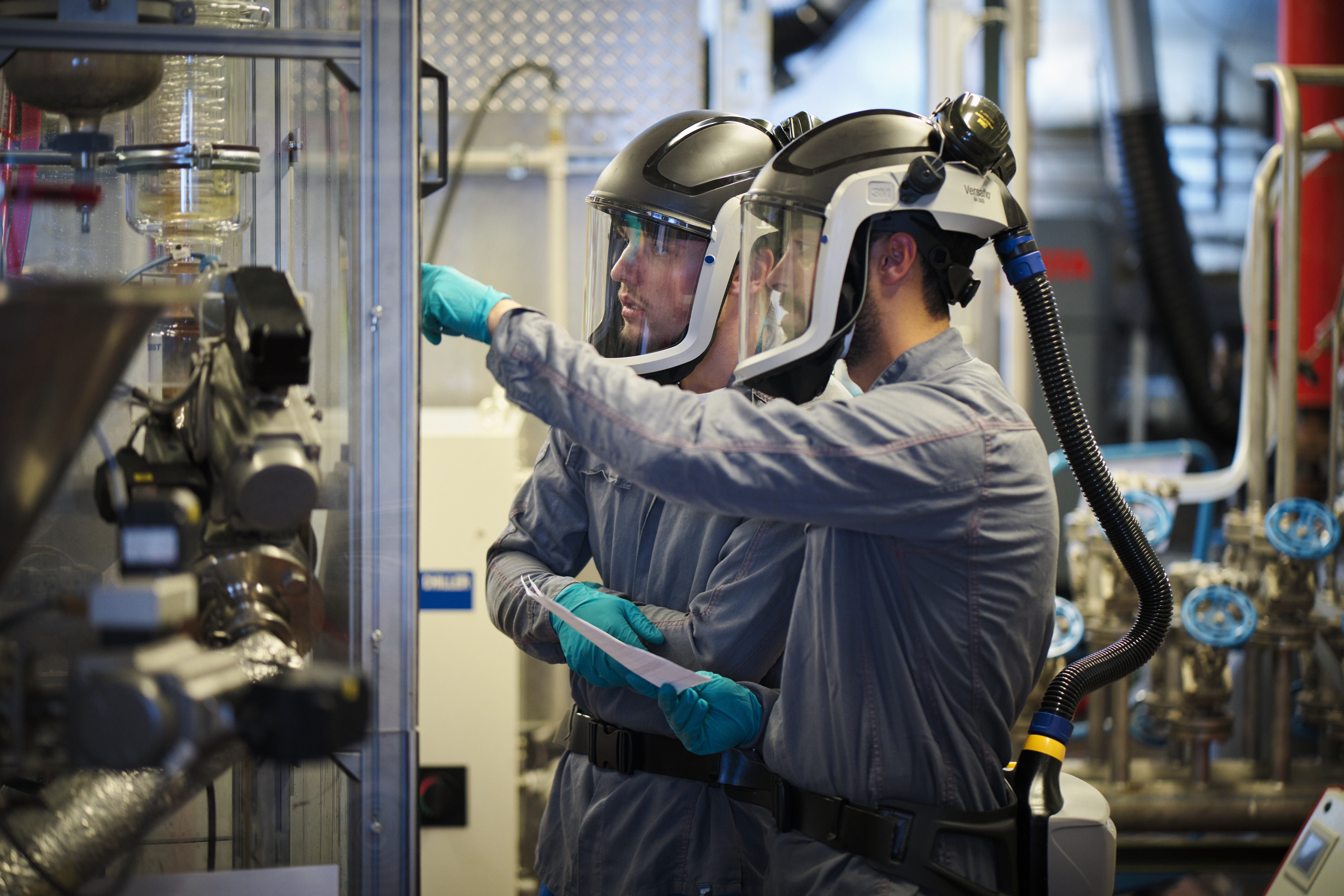 Two technicians in protective suits with powered air-purifying respirators inspect industrial chemical equipment.