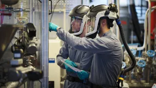 Two technicians in protective suits with powered air-purifying respirators inspect industrial chemical equipment.
