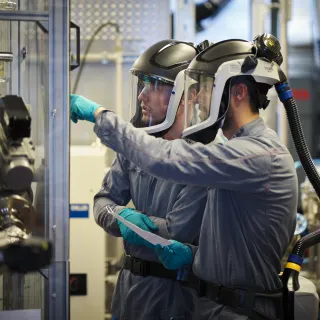 Two technicians in protective suits with powered air-purifying respirators inspect industrial chemical equipment.