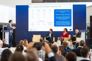 Speaker at a lectern on the left gesturing toward three seated panelists on stage, with a presentation slide behind them titled "M2i | Available Minerals & Metals – September 2026" showing tables of commodities.