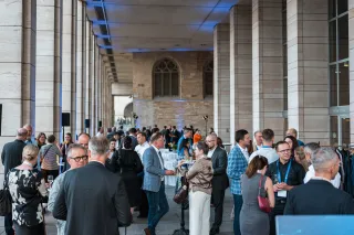 Conference attendees in business attire conversing and holding drinks under a high colonnade with stone pillars and historic brick arched windows in the background.